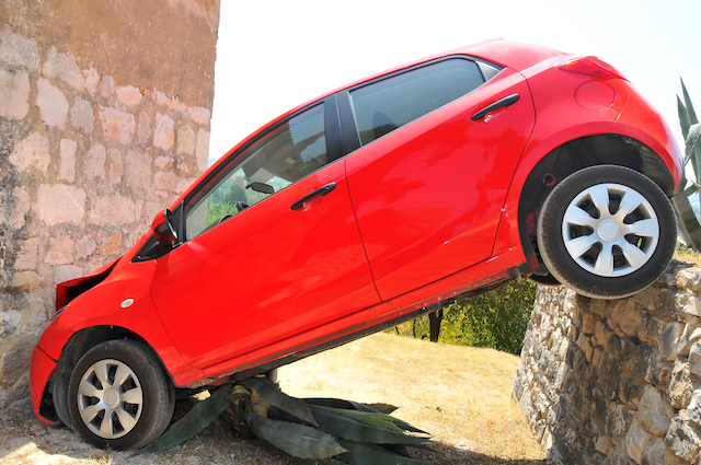 A small red car driven over a wall and crashed into the wall of a church, then wedged at an angle